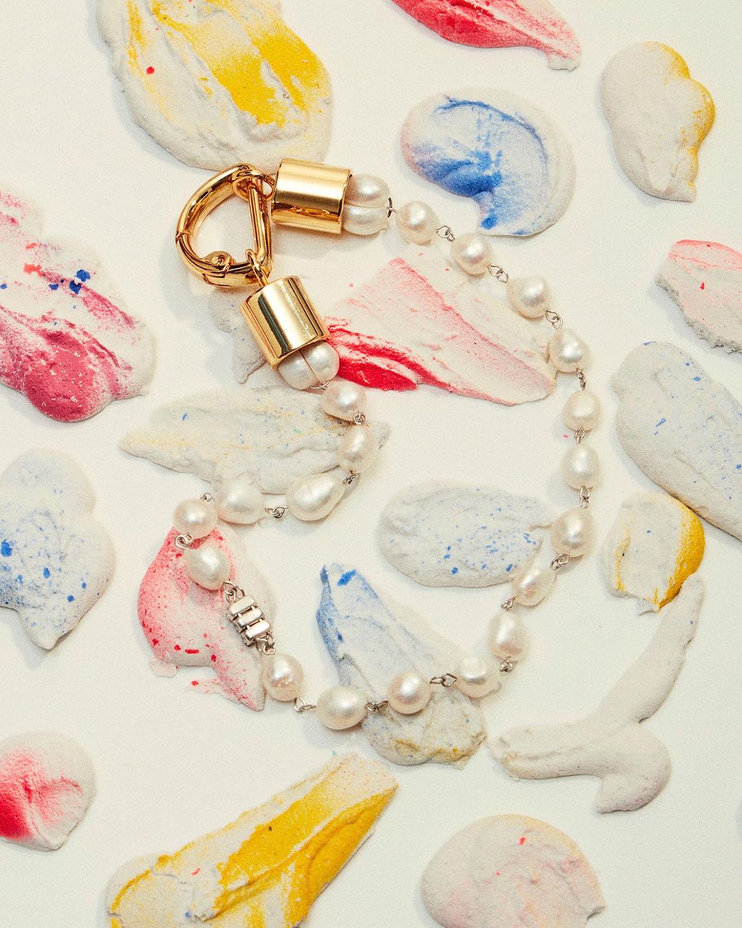 Overhead shot of a pearl necklace with gold accents against an abstract white background with red, blue, and yellow paint strokes.