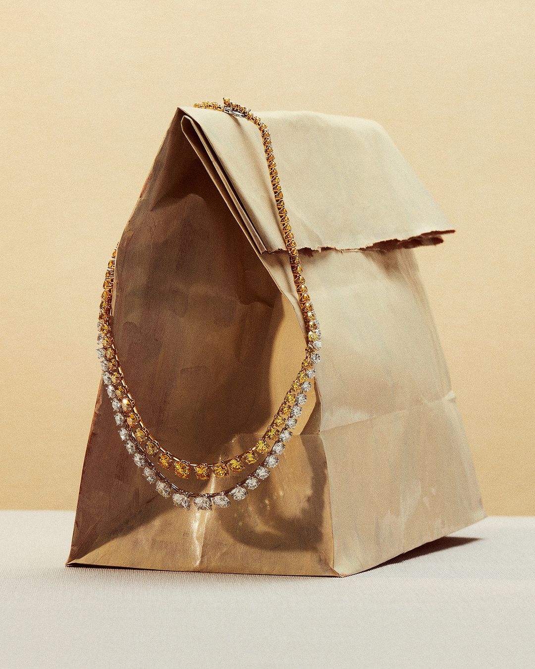 Gold and silver tennis necklace draped over a brown paper bag, studio shot for a jewelry product display, on a beige background.