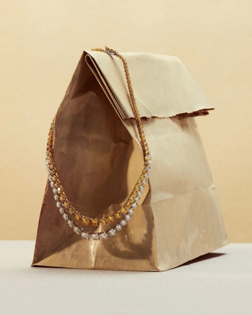 Gold and silver tennis necklace draped over a brown paper bag, studio shot for a jewelry product display, on a beige background.