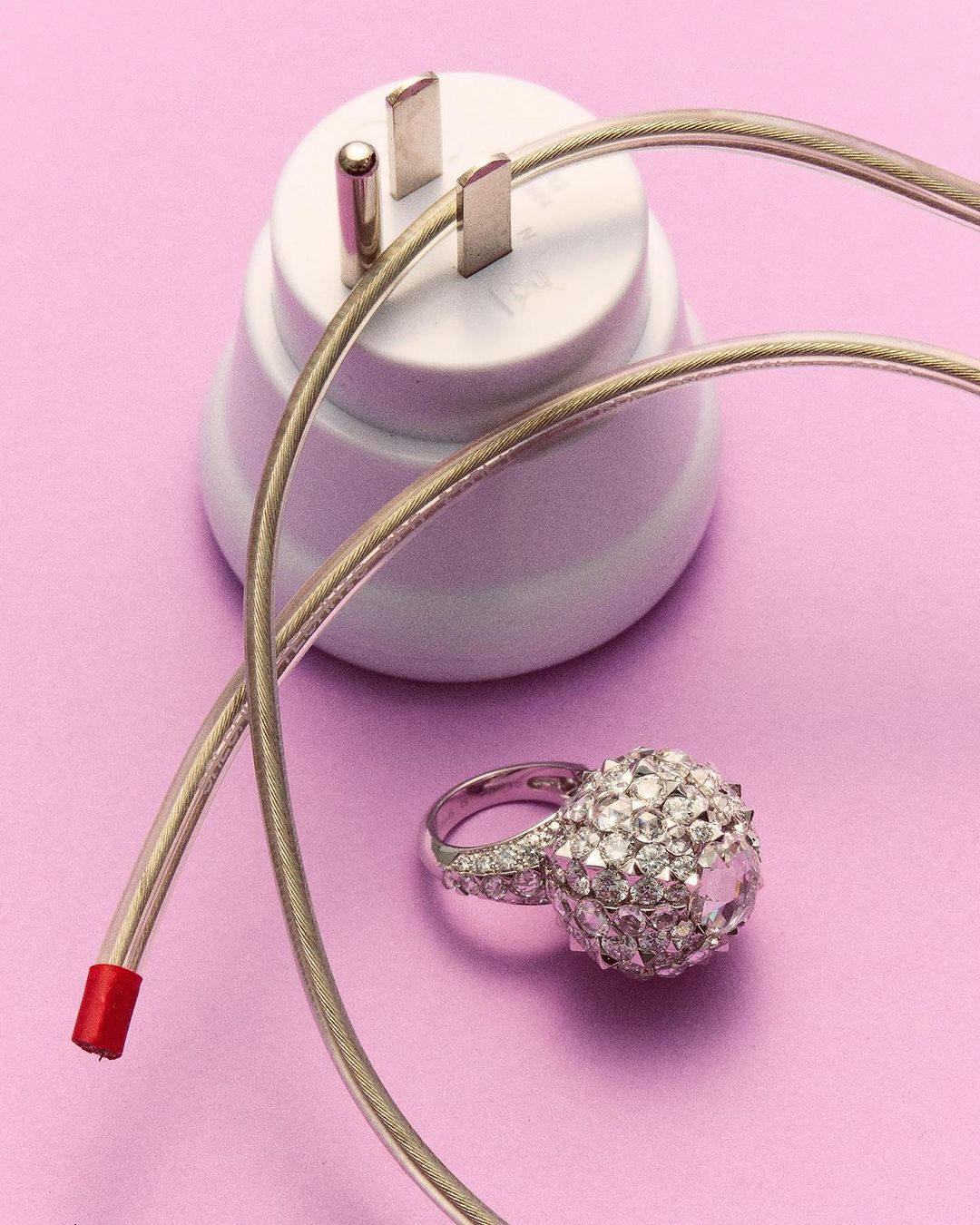 Still life shot of a jeweled ring beside an electrical plug and cord against a plain purple backdrop in a brightly lit studio setting.