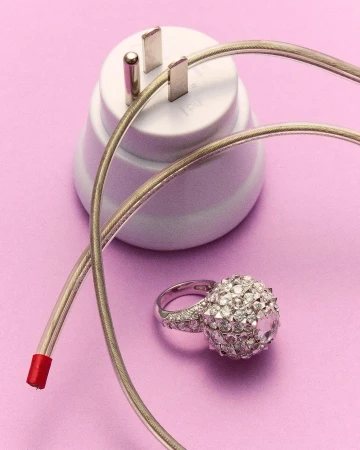 Still life shot of a jeweled ring beside an electrical plug and cord against a plain purple backdrop in a brightly lit studio setting.