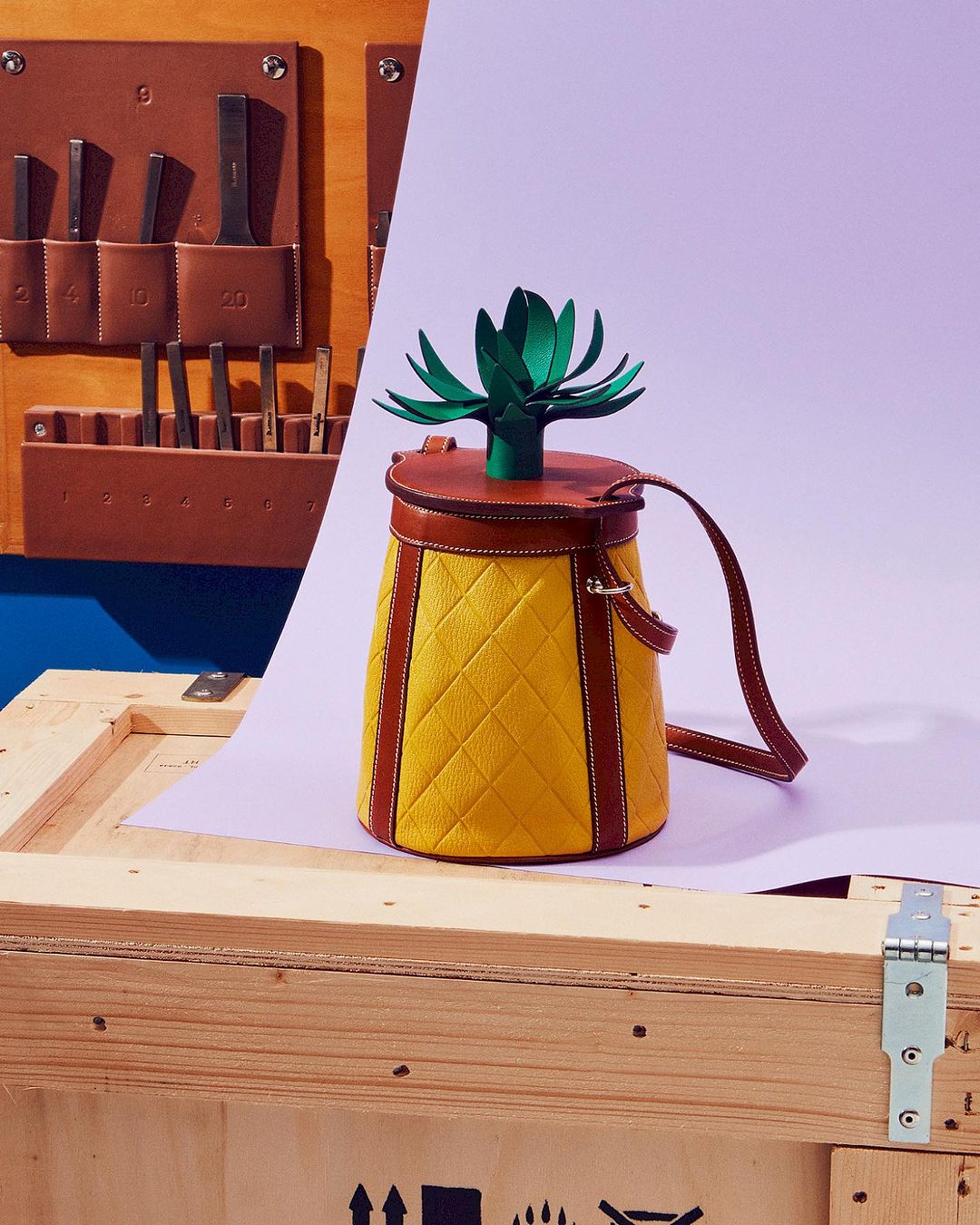 Pineapple-shaped handbag with yellow quilted texture, brown leather trim, and green paper foliage on a wooden crate against a purple background.