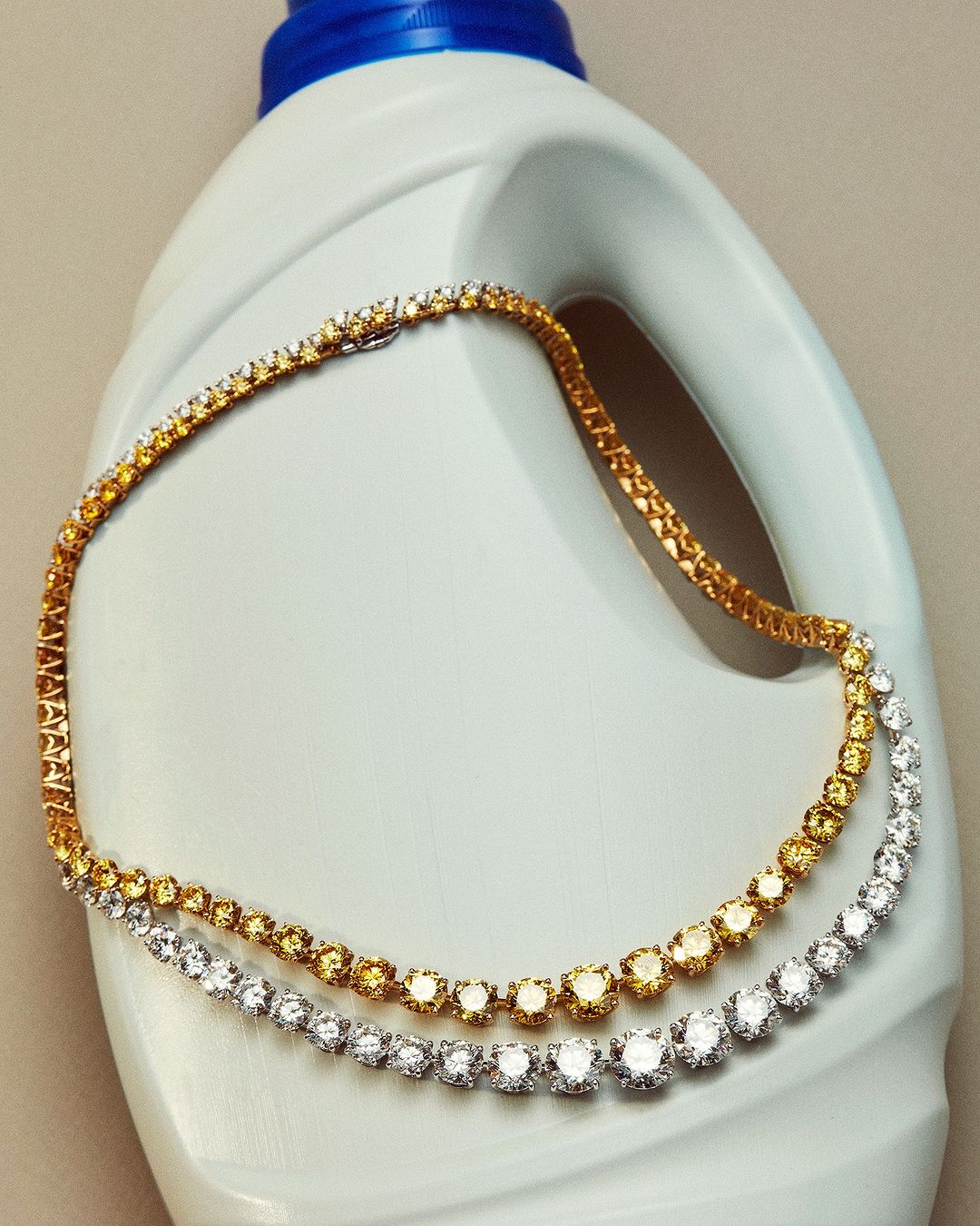 Two tennis necklaces, one gold and one silver, are draped across the top of a white and blue detergent bottle indoors with neutral background.