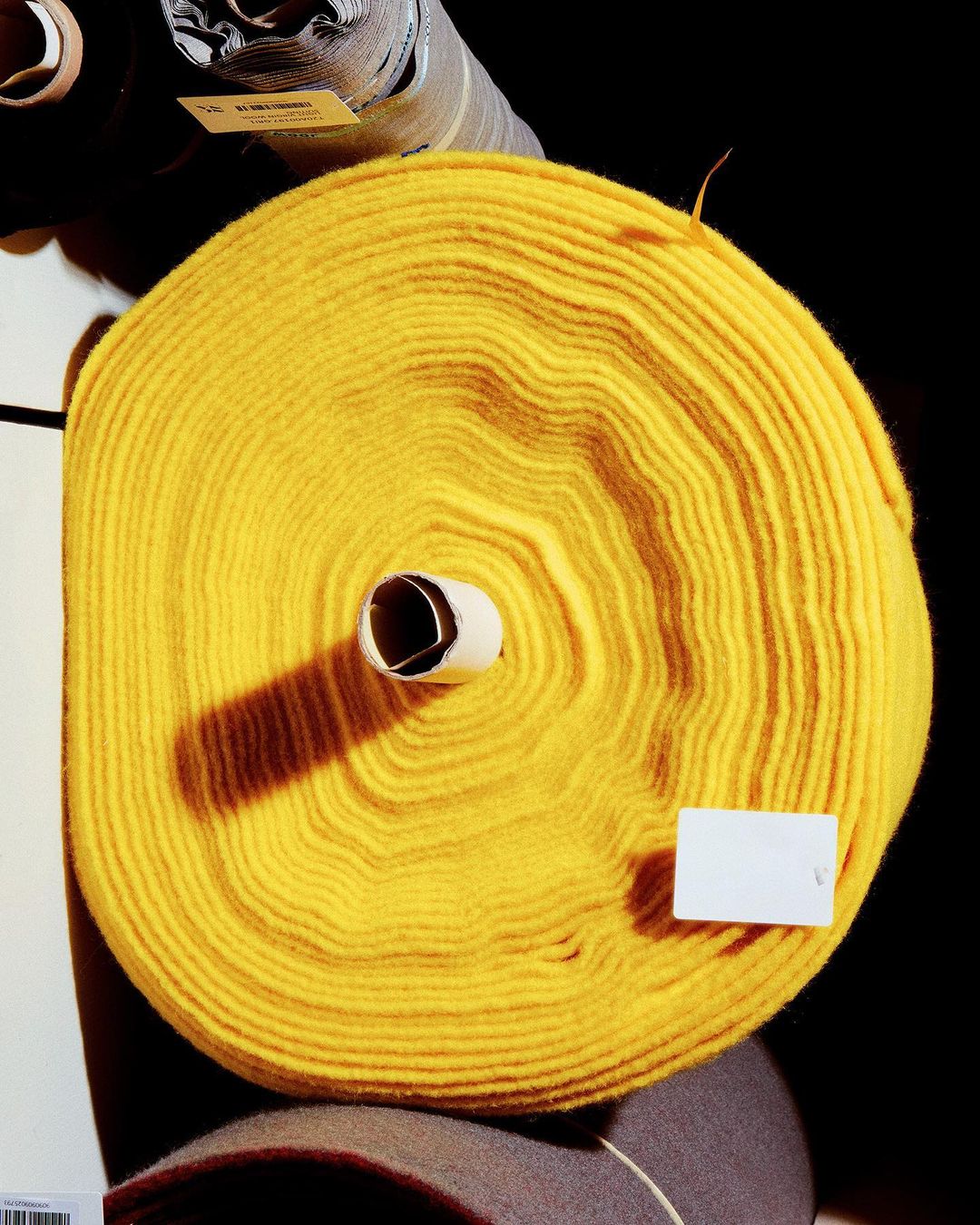 Close-up of a large roll of solid yellow fabric with paper core, labels, and other rolls of fabric visible in the background.
