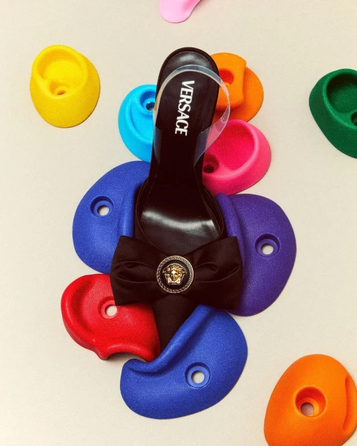 Elevated view of a black Versace high-heel shoe, adorned with a bow and Medusa head emblem, placed on colorful climbing holds.