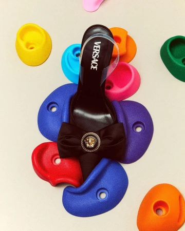Elevated view of a black Versace high-heel shoe, adorned with a bow and Medusa head emblem, placed on colorful climbing holds.
