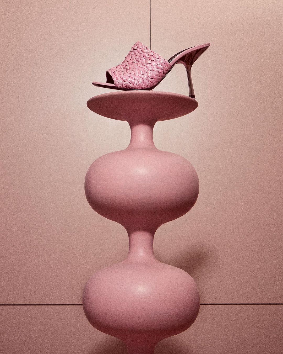 Pink woven mule heeled sandal on a pink abstract sculpture in a pink room creates a minimalist fashion product still life.