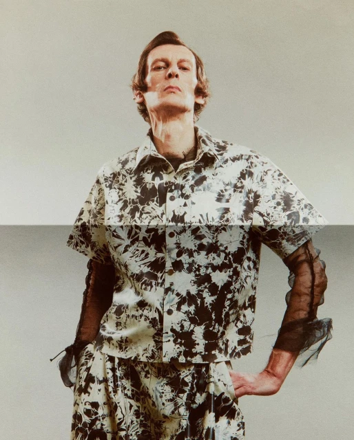 A light-skinned man in a patterned shirt and shorts poses in a studio with a neutral background. He stands with one hand on his hip.