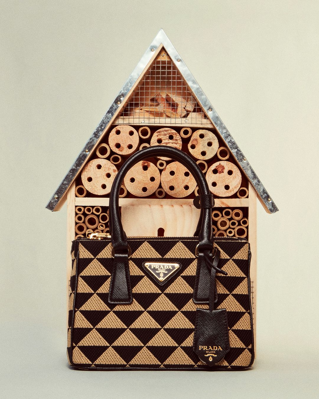 Prada handbag in front of a wooden insect hotel, set against a neutral background. The bag features black triangular patterns.