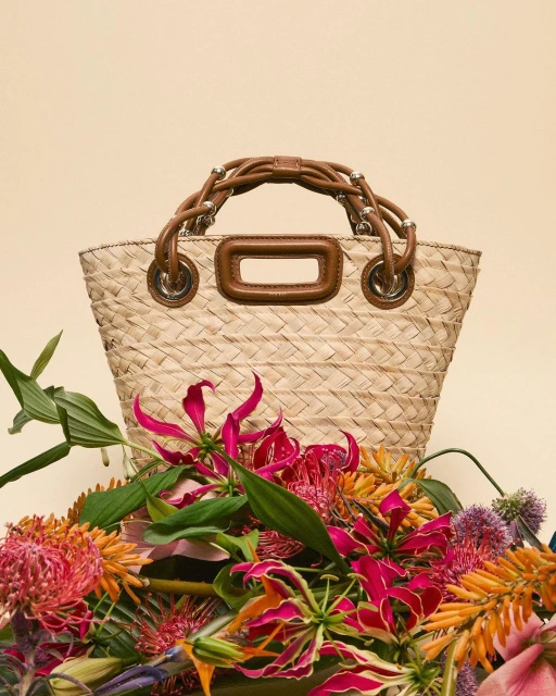 Woven straw bag with brown leather handle, encircled by vibrant flowers against a neutral backdrop, creating a still-life fashion shot.