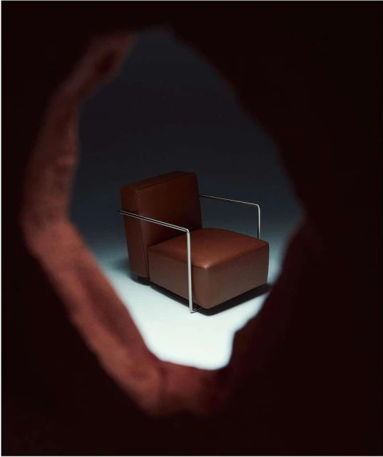 Brown leather armchair with metal frame, presented in a spotlight through a textured hole, creating an intimate and focused composition.