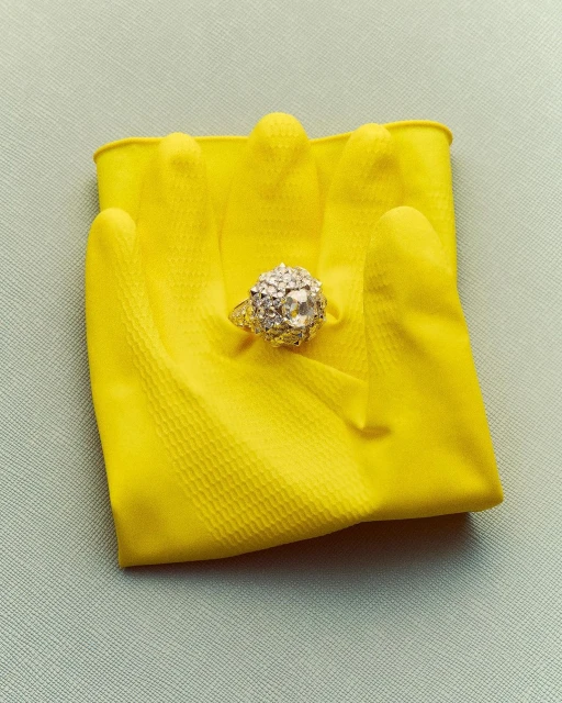 Engagement ring on a yellow rubber glove, placed on a light blue background. The ring is large, detailed, and diamond-encrusted.