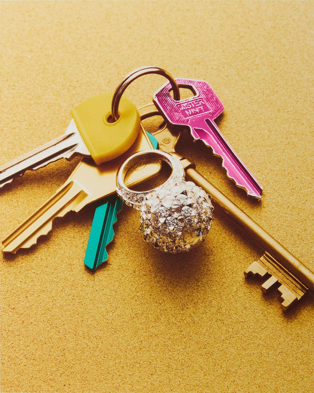 A collection of colorful metal keys and a sparkling ring arranged on a textured gold surface in a bright, eye-level studio shot.