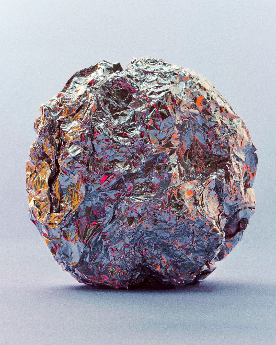 Shiny silver ball made of crumpled aluminum foil, reflecting shades of orange, pink, and blue, against a soft grey background.