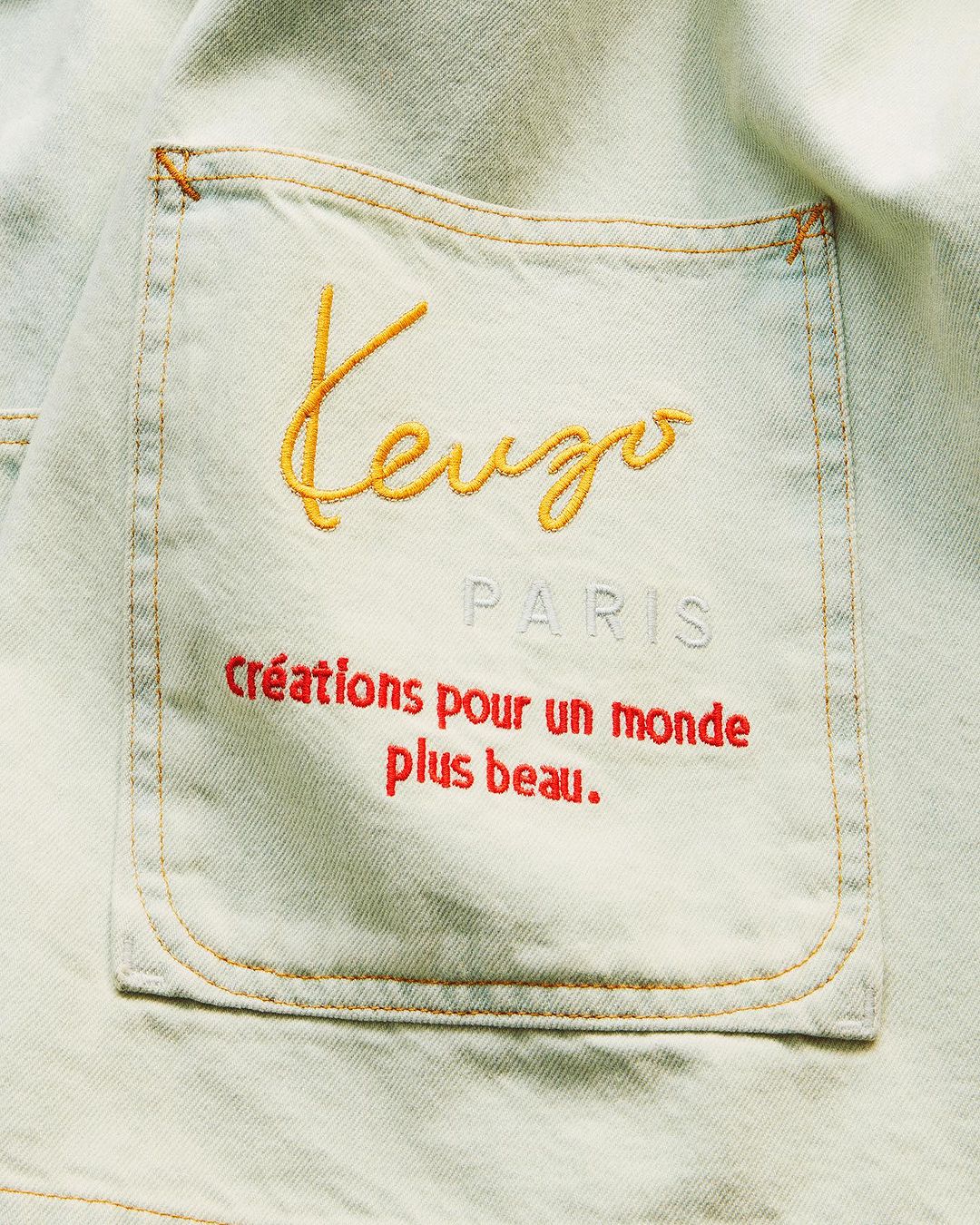 Close-up of a light blue denim jacket with an embroidered cream-colored Kenzo logo, 'Paris' in gray, and red French text.