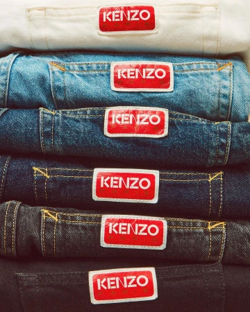 Close-up of folded denim jeans with Kenzo brand labels, stacked in a variety of colors, displaying a range of blue hues and textures.