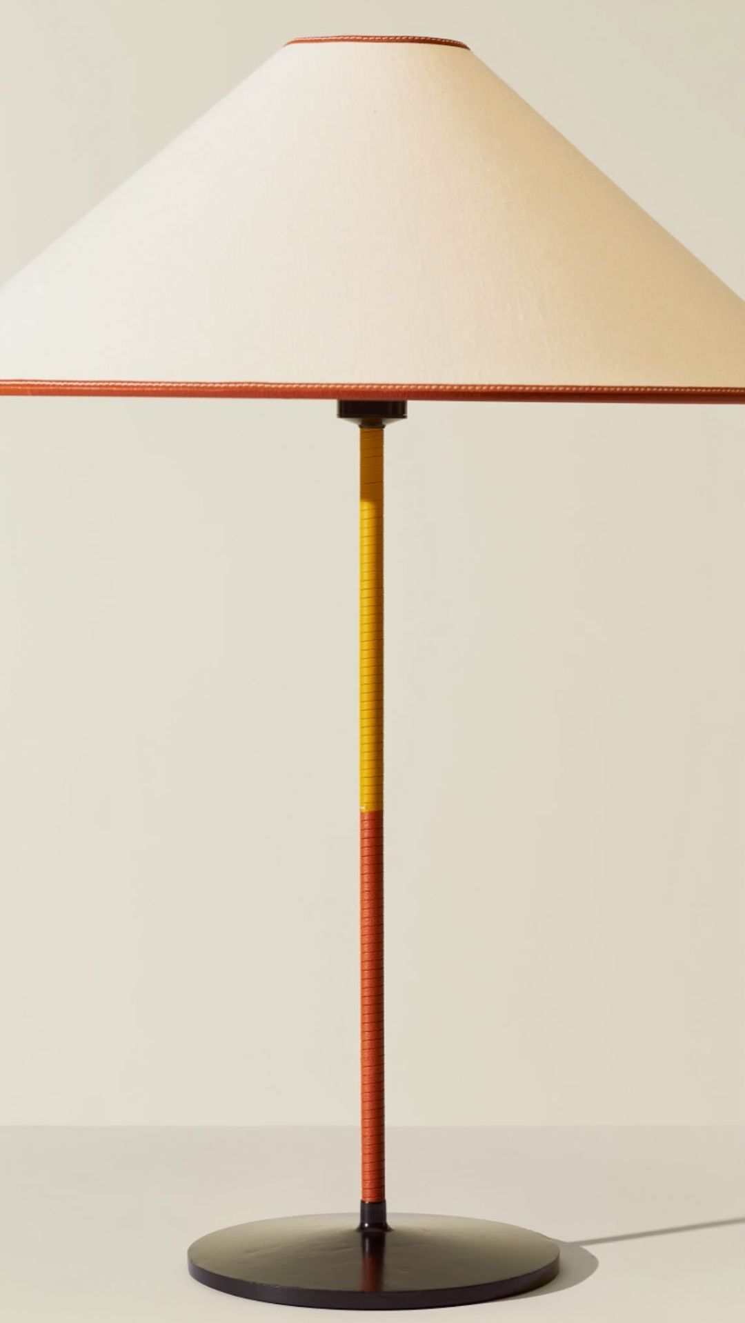 Minimalist table lamp featuring a conical cream shade with tan trim and a two-tone yellow and tan stand on a circular black base.