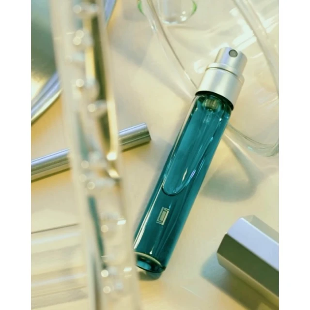 Turquoise perfume spray bottle with silver cap on a white reflective surface, surrounded by clear glass containers and cylinder.