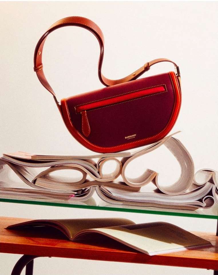 Burgundy Burberry shoulder bag with brown leather strap on clear shelf with stack of magazines and wood shelf against white background.