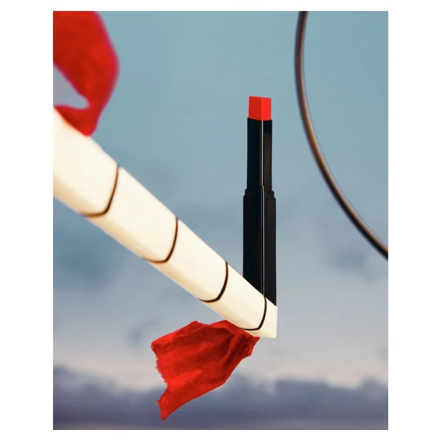 Close-up studio shot of a red lipstick tube, diagonally positioned with a fabric accent, against a soft blue, cloudy sky background.