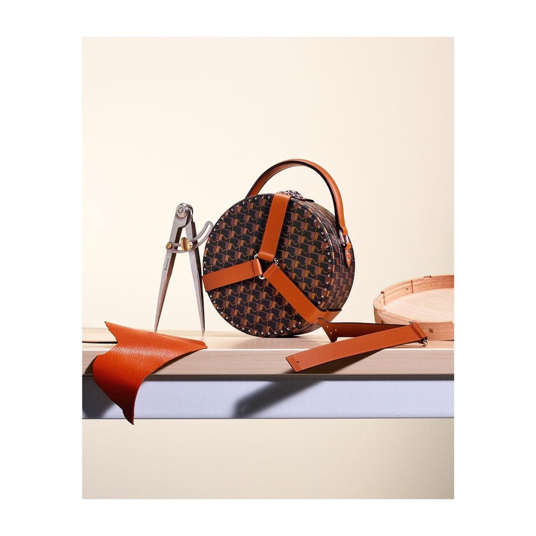Round handbag with pattern and leather straps on wooden surface, along with leather scraps and a tool, in a studio setting.