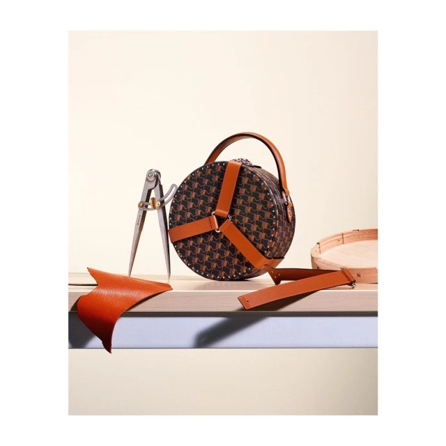Round handbag with pattern and leather straps on wooden surface, along with leather scraps and a tool, in a studio setting.