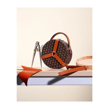 Round handbag with pattern and leather straps on wooden surface, along with leather scraps and a tool, in a studio setting.