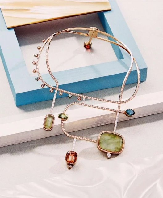 Elegant gold necklace featuring green and multi-colored gemstones displayed on a blue surface with a white backdrop.