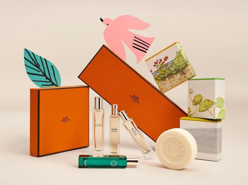 Hermès perfume bottles and product boxes arranged with paper bird and leaf props in a beauty product still life on a cream background.