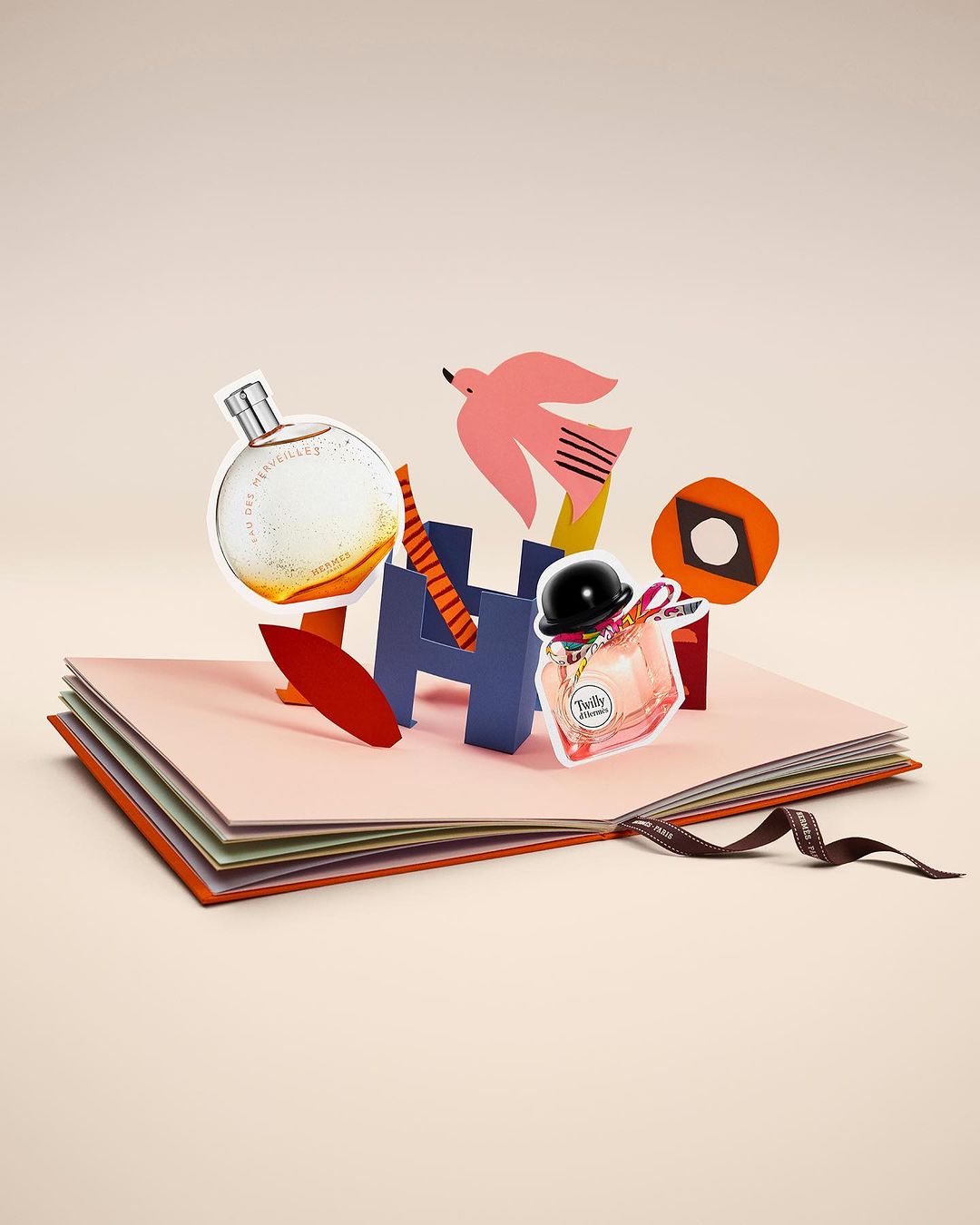 Still life of perfume bottles displayed on a paper pop-up book with paper cut-out decorations against a light peach background.