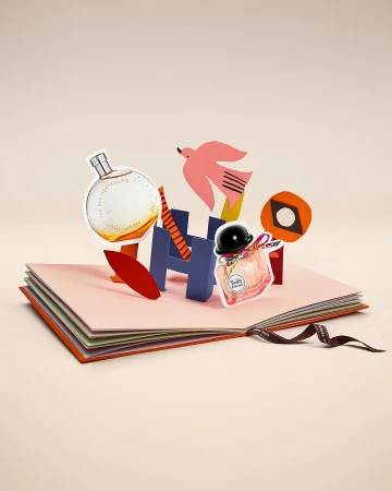 Still life of perfume bottles displayed on a paper pop-up book with paper cut-out decorations against a light peach background.