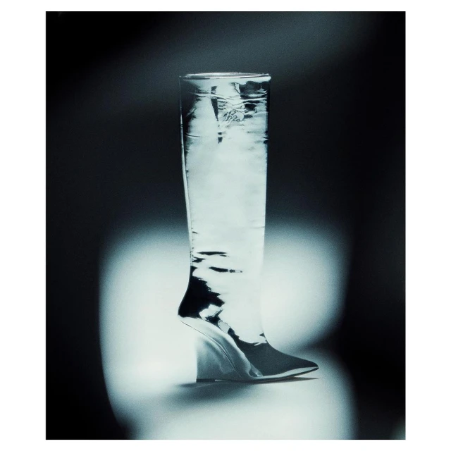 High heel boot made of glass, lit with a spotlight, against a dark backdrop. The clear material allows the light to shine through.