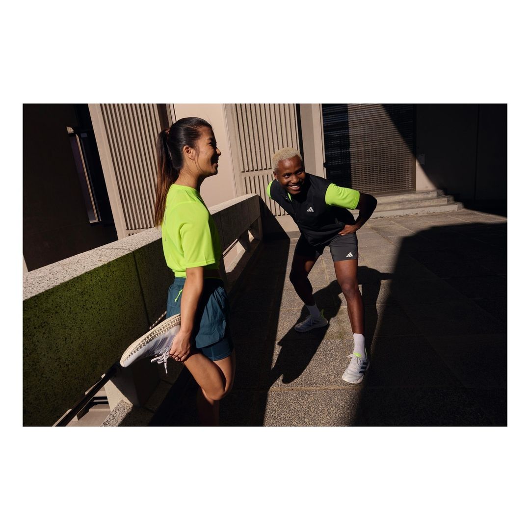 Two athletes, an Asian woman in neon green and a Black man in black and neon green, stretch together on an outdoor urban walkway.