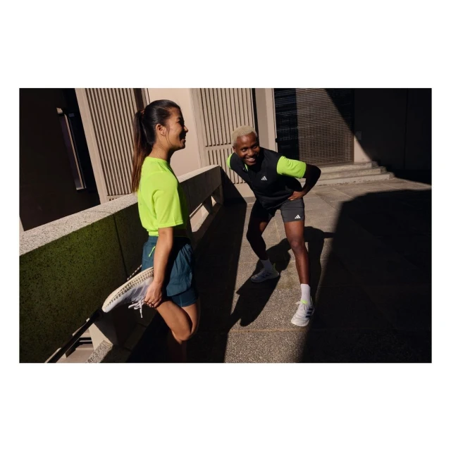 Two athletes, an Asian woman in neon green and a Black man in black and neon green, stretch together on an outdoor urban walkway.