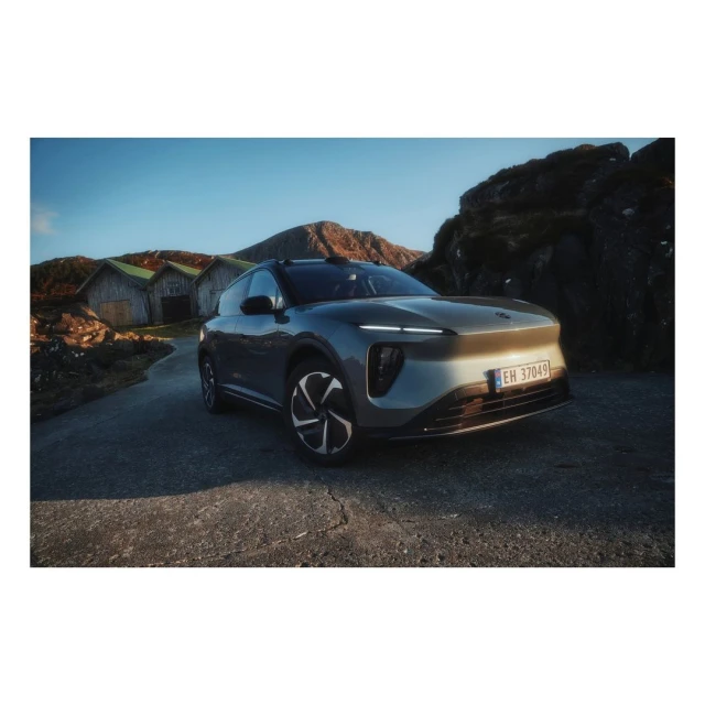 Grey NIO SUV parked on a cracked asphalt road in front of wooden houses and rocky hills beneath a clear blue sky on a sunny day.