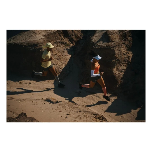 Two athletic adults run across a sandy terrain in bright sunlight. The runners wear athletic apparel and hats in outdoor scene.