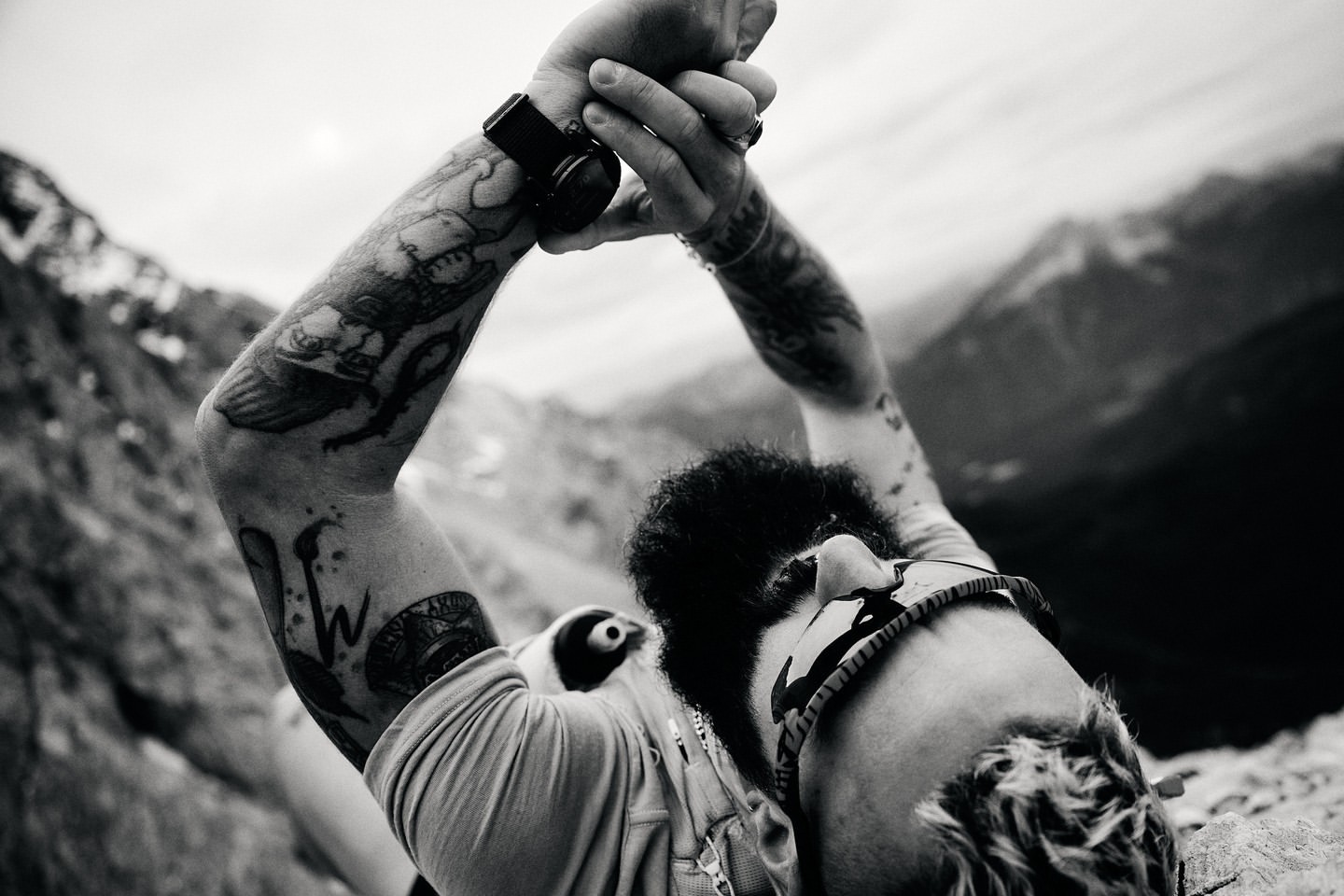 Close-up monochrome portrait of a tattooed man with a beard wearing sunglasses and checking his watch outdoors.
