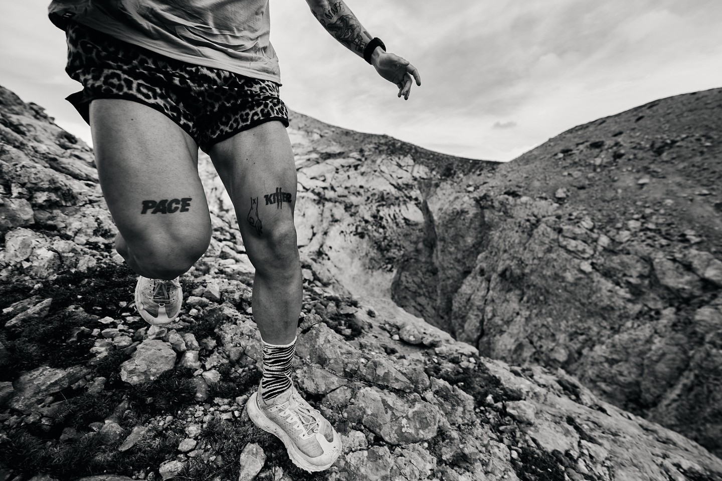 Monochrome action shot of a male runner in motion on a rocky mountain trail with tattoos and patterned shorts, running shoes, and socks.