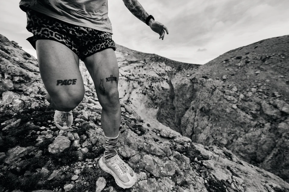 Monochrome action shot of a male runner in motion on a rocky mountain trail with tattoos and patterned shorts, running shoes, and socks.