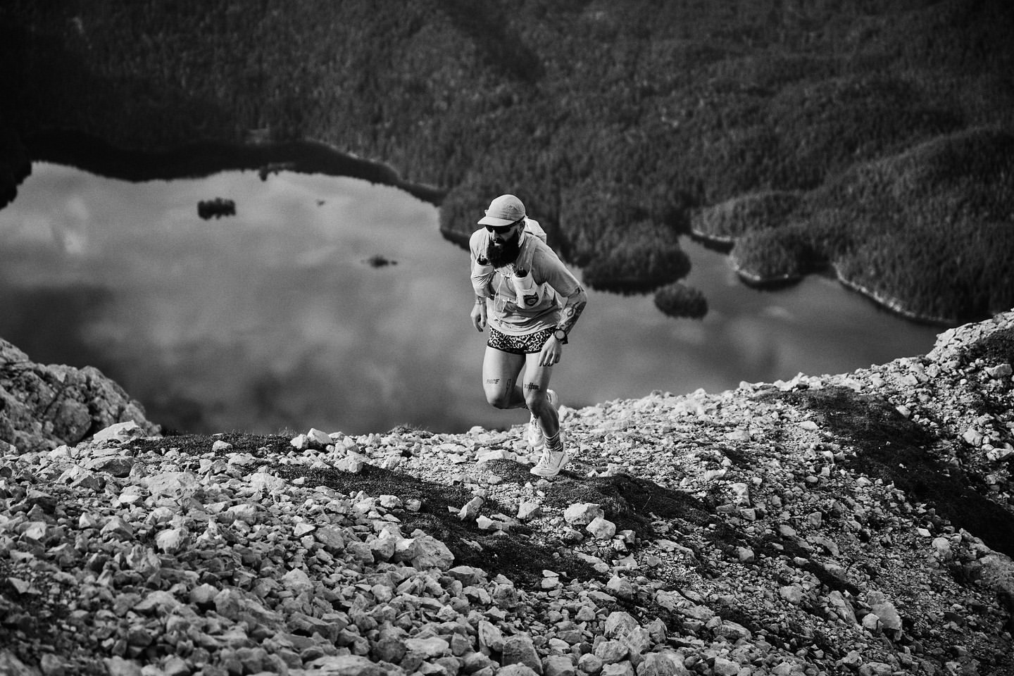 Bearded man trail running uphill on a rocky landscape with mountains and a lake in the background in a black and white outdoor shot.
