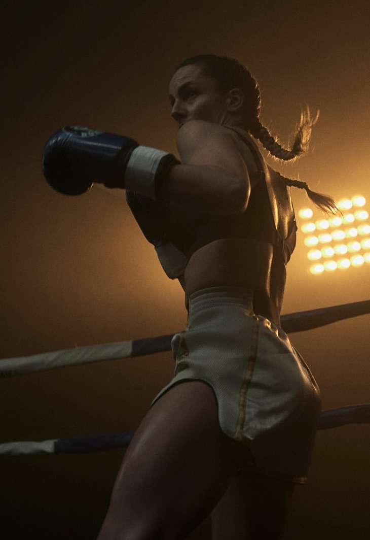 Female boxer in ring. Athlete in boxing stance with blue gloves, wearing white boxing shorts and sports bra, in a boxing arena.