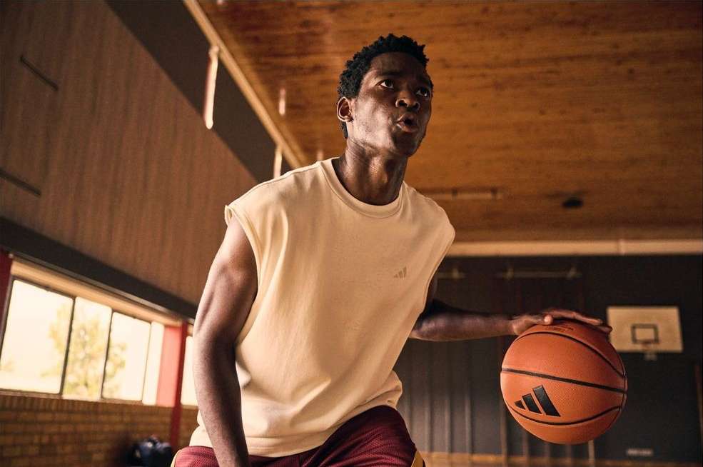 An African-American man dribbles an Adidas basketball on an indoor court with a focused expression, wearing a yellow t-shirt and red shorts.