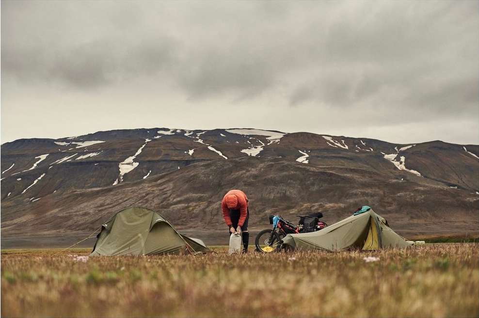 Person inflating camping gear near a bicycle and tents in a grassy field with a mountain backdrop under an overcast sky.