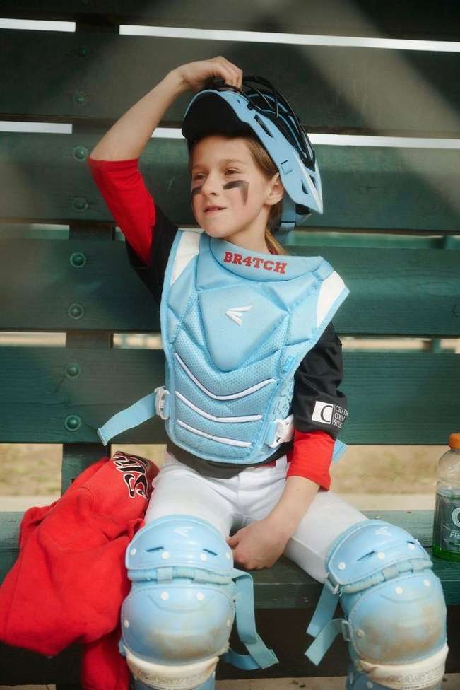 Young baseball player in full protective gear, including a helmet and chest protector, sits on a bench after a game.