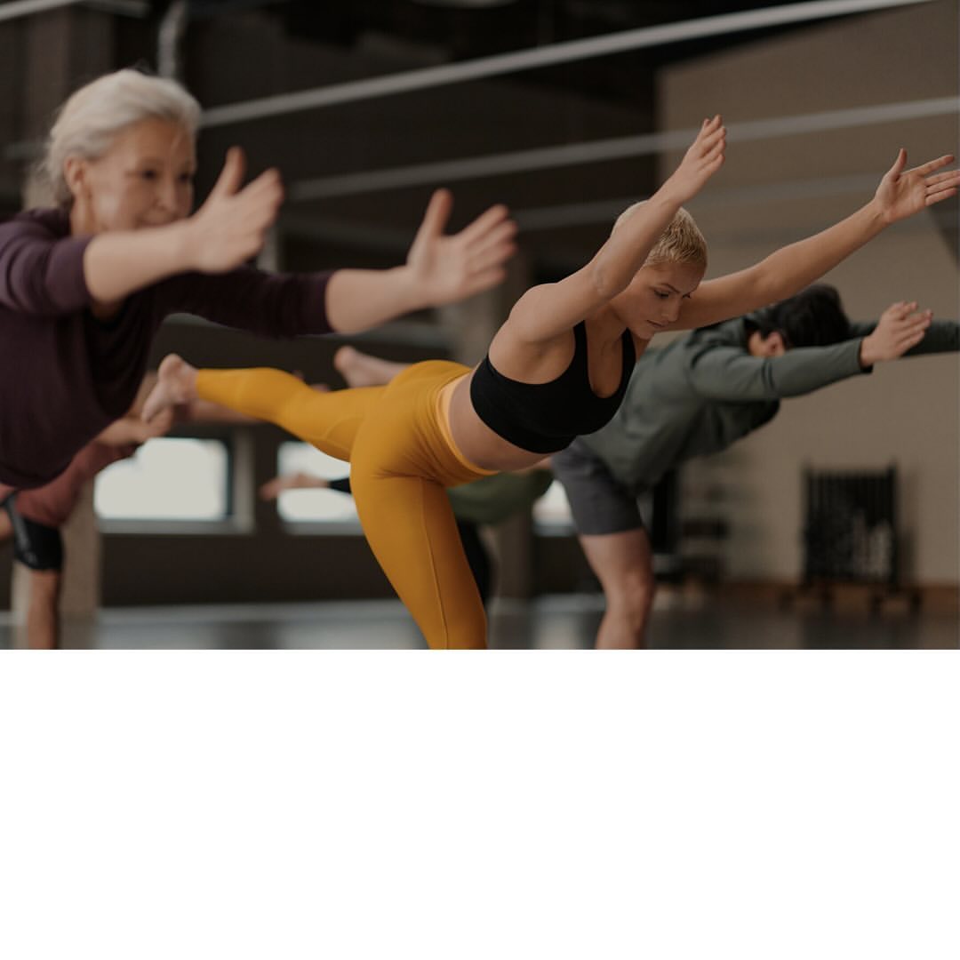 Group of people in a yoga studio practicing balancing poses. Blond woman in a black sports bra and yellow leggings is in the foreground.