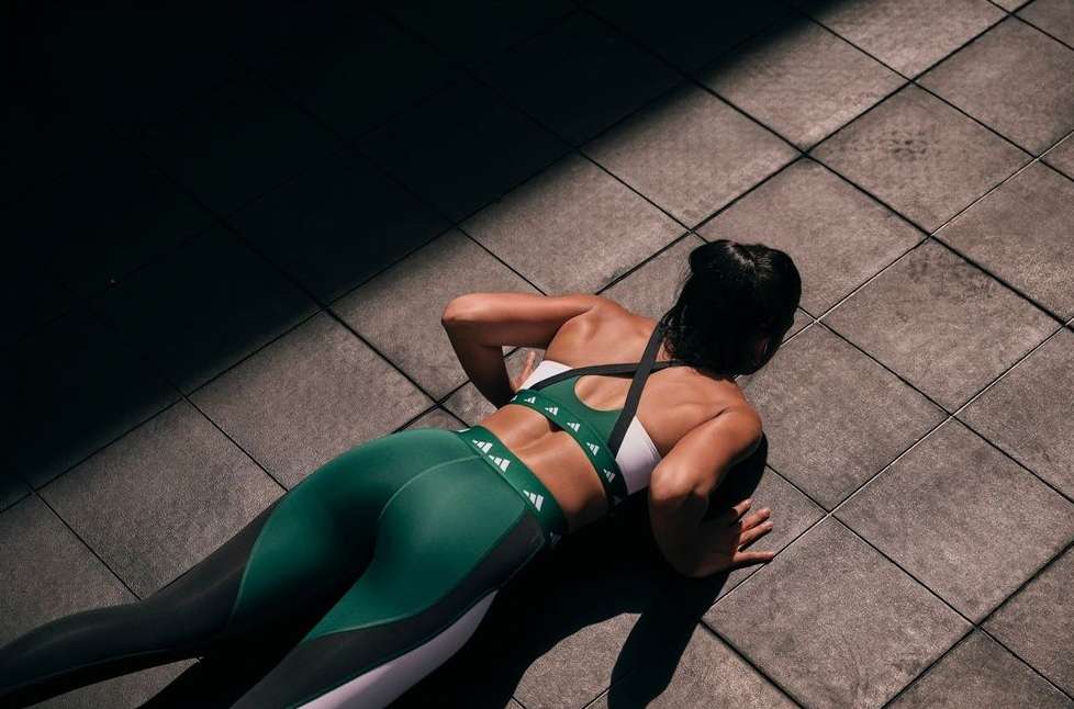 From a back view, an adult woman in fitness apparel does push-ups on a gray tiled floor with a dark shadow on the upper left.