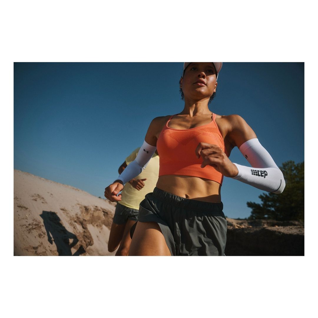 Two runners, one adult female in the foreground, wearing athletic wear, and arm sleeves, running on a sunny day in nature.