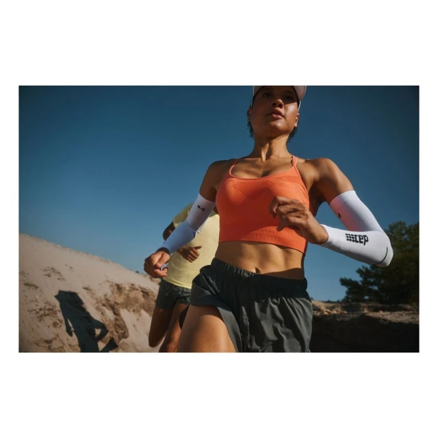 Two runners, one adult female in the foreground, wearing athletic wear, and arm sleeves, running on a sunny day in nature.
