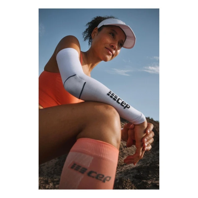 Woman in fitness apparel poses outdoors in sun. She is wearing a visor, arm sleeves, athletic top and compression socks against blue sky.