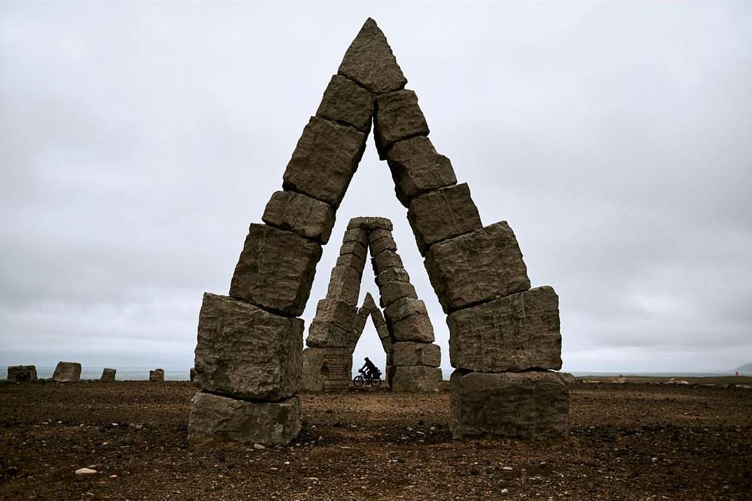 Person on a bike riding through a stone archway on a cloudy day, low angle shot. The archway is made of stacked stone blocks.